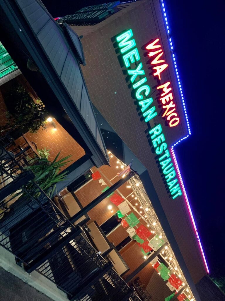 Outdoor view of Viva Mexico Mexican Restaurant at night, featuring neon signs and a patio decorated with colorful string banners.