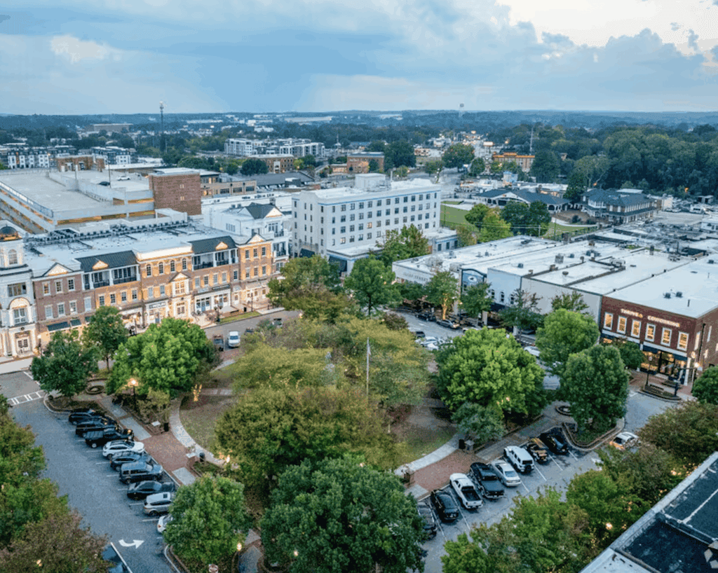 Aerial view of a small town square with parked cars, surrounding trees, historic-style buildings, and a partly cloudy sky—ideal spot for a turnkey restaurant for sale.