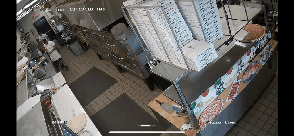 Surveillance camera view of a profitable pizzeria kitchen in Dawsonville GA, showing stacked pizza boxes, prep counters, and a person working in the background.
