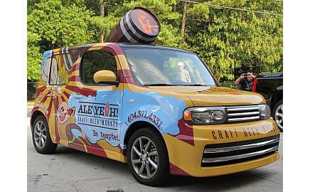 A yellow car with colorful craft beer-themed graphics and a large barrel decoration on its roof, parked outside a Decatur GA Craft Beer Store, hints at this profitable business opportunity surrounded by trees.